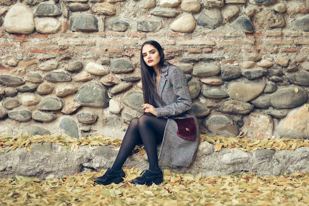 Muchacha hermosa joven con el abrigo de invierno que lleva del pelo muy largo que se sienta en el piso de un parque urbano