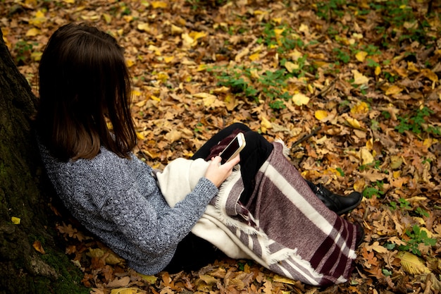 Muchacha hermosa joven en una chaqueta gris que se sienta en el bosque del otoño cerca de un árbol grande con un teléfono móvil