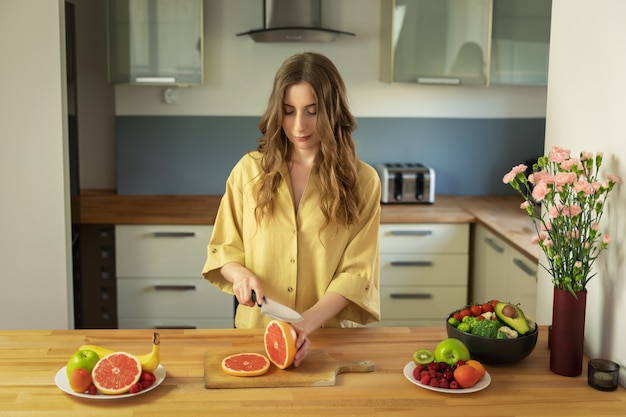 La muchacha hermosa joven está cortando una toronja. Una mujer prepara una ensalada de frutas deliciosas y saludables.