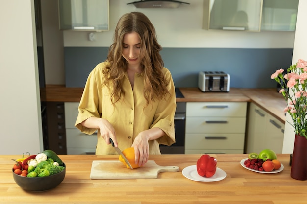 La muchacha hermosa joven con el pelo largo corta la pimienta dulce. Una mujer prepara una ensalada de verduras frescas y saludables.