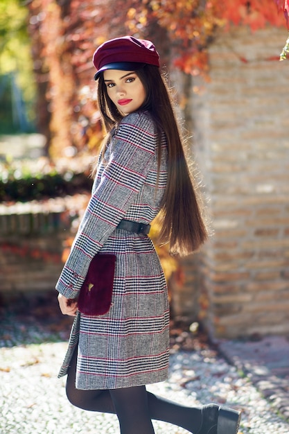 Muchacha hermosa joven con el pelo muy largo que lleva el abrigo de invierno y el casquillo en hojas de otoño