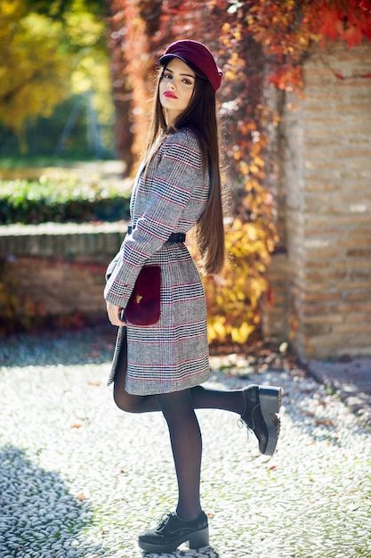 Muchacha hermosa joven que lleva el abrigo de invierno y el casquillo en fondo de las hojas de otoño.