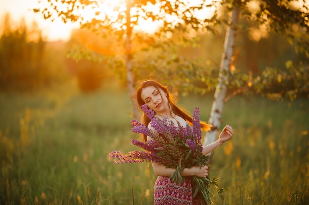 Muchacha hermosa joven que sostiene una flor grande con lupine púrpura en un campo floreciente. Blooming flores de lupino.