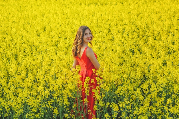 La muchacha hermosa joven en un vestido rojo se cierra para arriba en el medio del campo amarillo con las flores del rábano. Temporada de primavera