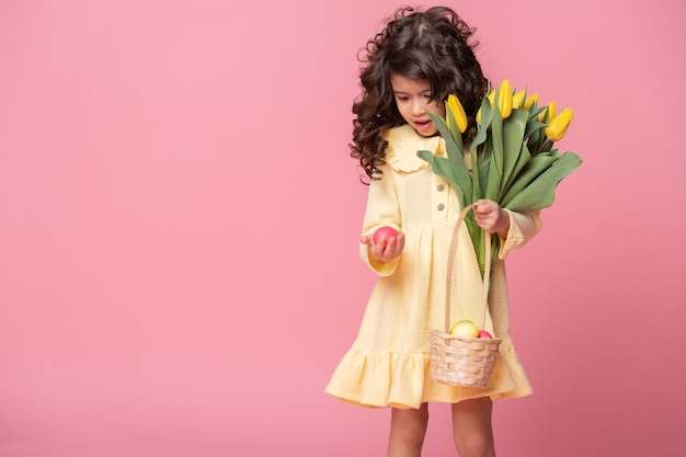 Muchacha hermosa del niño que sostiene la cesta con los huevos de Pascua y los tulipanes en fondo rosado.