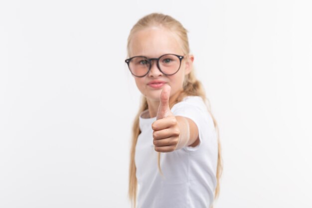 Muchacha hermosa del niño con los vidrios que muestran los pulgares para arriba aislados en blanco