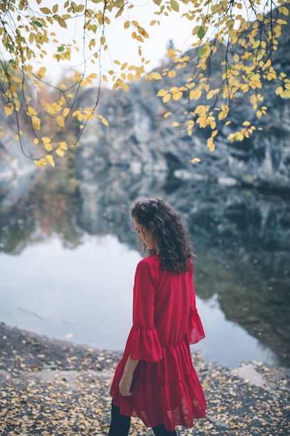 Muchacha hermosa del pelo rizado en un vestido rojo que camina por el lago.