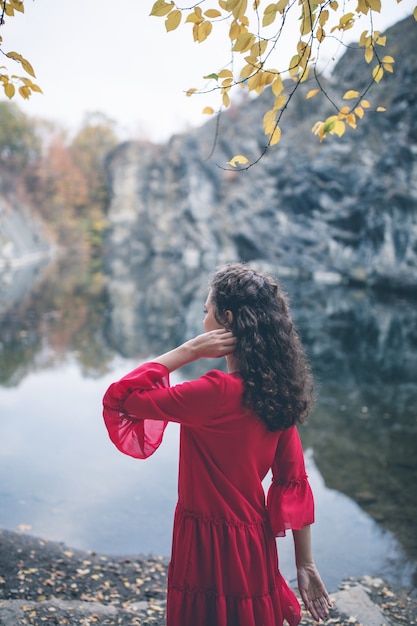 Muchacha hermosa del pelo rizado en un vestido rojo que mira el lago