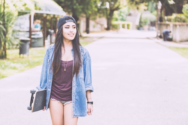 Muchacha hermosa que camina en el parque que sostiene un monopatín.