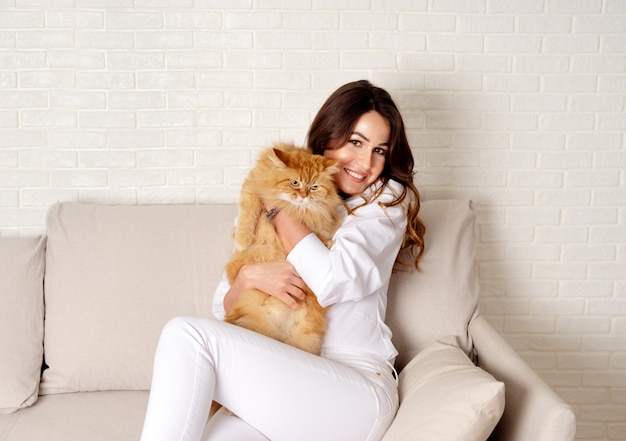 Muchacha hermosa que se sienta con un gato rojo mullido en el sofá