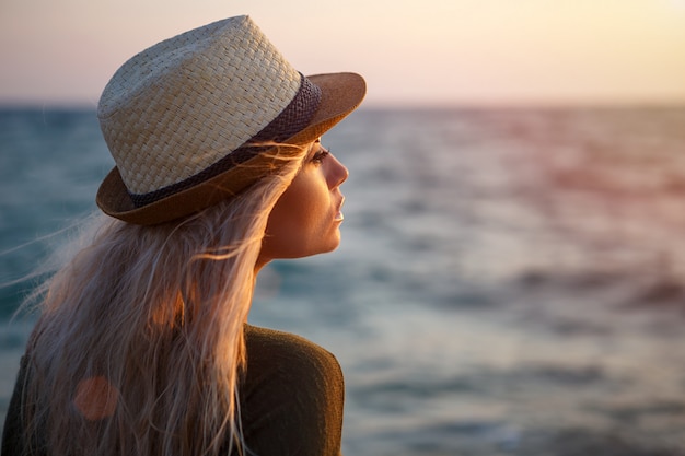 Muchacha hermosa en sombrero por el mar en la puesta del sol.