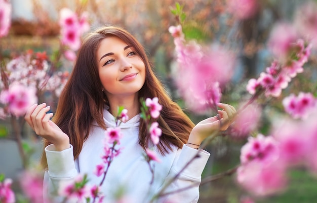 Muchacha hermosa sonriente que se coloca cerca de un árbol de melocotón durante puesta del sol. Cara feliz. Tiempo de primavera.