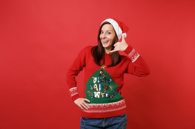 La muchacha joven alegre de Santa que hace el gesto del teléfono como dice: devuélvame la llamada con la mano, los dedos como hablando por teléfono aislado sobre fondo rojo. Feliz año nuevo 2019 celebración concepto de fiesta navideña.