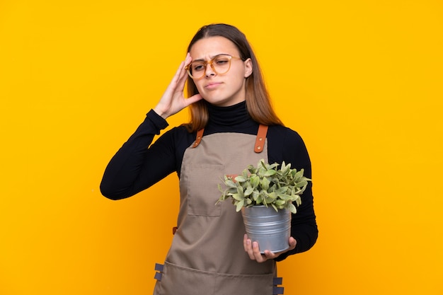 Muchacha joven del jardinero que sostiene una planta sobre amarillo aislado infeliz y frustrado con algo. Expresión facial negativa