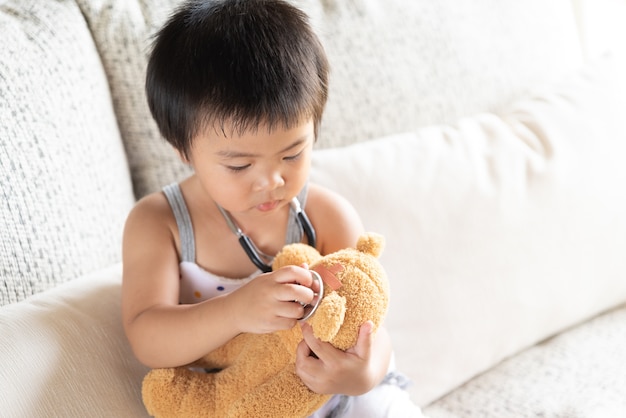 La muchacha linda está jugando al doctor con el estetoscopio y el oso de peluche.