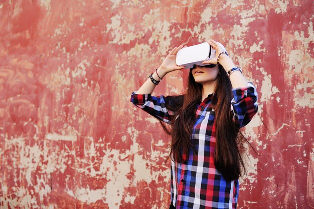 Muchacha linda joven con el pelo largo en camisa de tela escocesa en vidrios de la realidad virtual en fondo rojo de la pared del grunge.