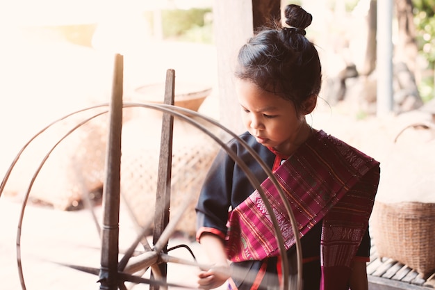 Muchacha linda del niño asiático en el vestido tradicional tailandés que usa la rueda de giro de madera tradicional