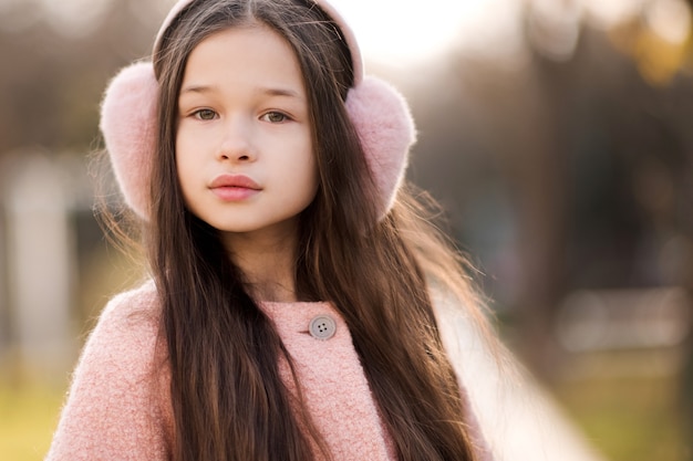 Muchacha linda del niño del estilo que lleva los auriculares y la chaqueta mullidos al aire libre sobre la naturaleza