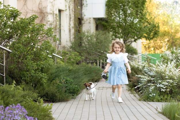 Muchacha linda del niño en el vestido que lleva al perro pequeño a pasear al aire libre en la naturaleza. Amistad entre los animales y los niños.