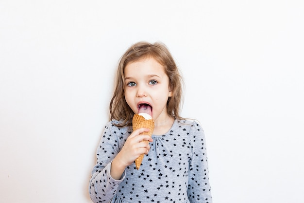 Muchacha linda que come el helado que presenta sobre la pared blanca. ¡Hora de verano!