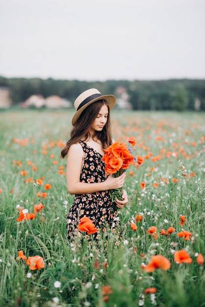 La muchacha modelo morena joven hermosa en un sombrero camina en un campo de flores con un ramo de amapolas. Campo fragante de flores rojas.