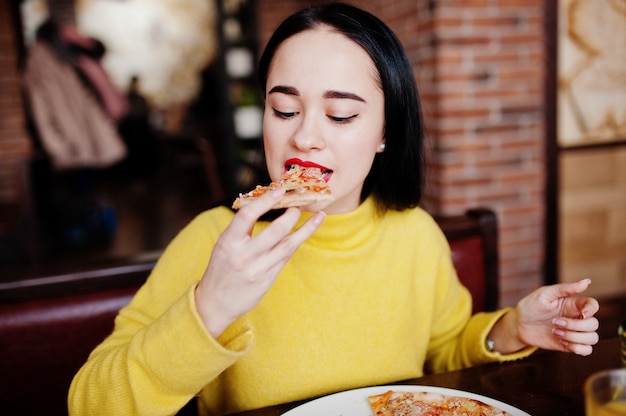 Muchacha morena divertida en suéter amarillo que come la pizza en el restaurante.