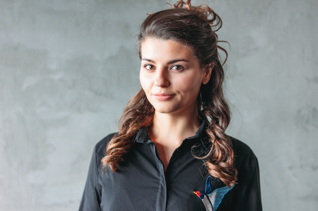 Muchacha morena sonriente sonriente joven con el pelo ondulado en el vestido negro de la camisa, aislado en gris