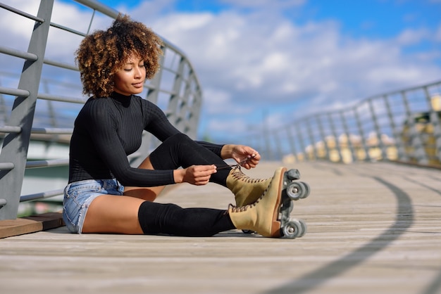 La muchacha negra sonriente de los jóvenes que se sienta en el puente urbano y pone patines.