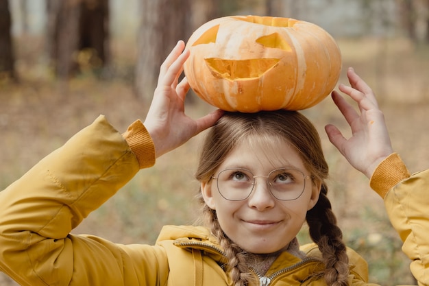 Muchacha del niño feliz con calabaza de Halloween en la cabeza. Preparación para la fiesta en el jardín cerca de las decoraciones de Jack-o-Lantern