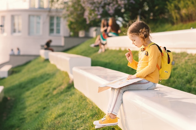 Muchacha del niño en ropa amarilla con el libro de lectura de la mochila en lugar de la tumbona en el parque al aire libre soleado en verano. Regreso a la escuela.