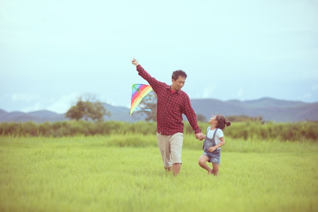 Muchacha y padre del niño asiático con una cometa que corre y feliz en prado en verano en naturaleza