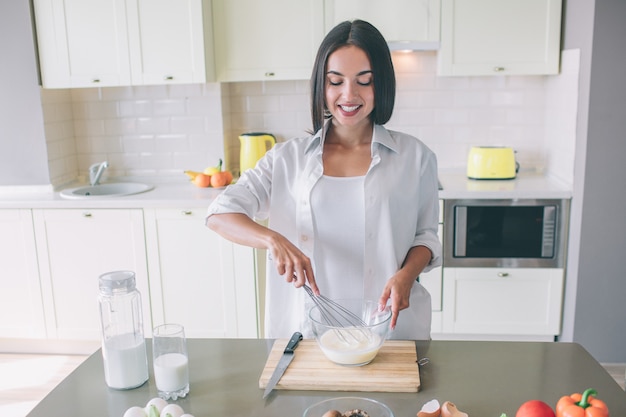 La muchacha positiva y agradable se coloca en la cocina en la mesa y mezcla la leche con los huevos.