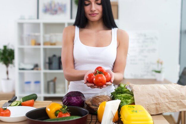 Muchacha que se coloca en la cocina que sostiene el manojo rojo del tomate en sus manos. Ama de casa tomando ingredientes frescos para ensalada de verduras.