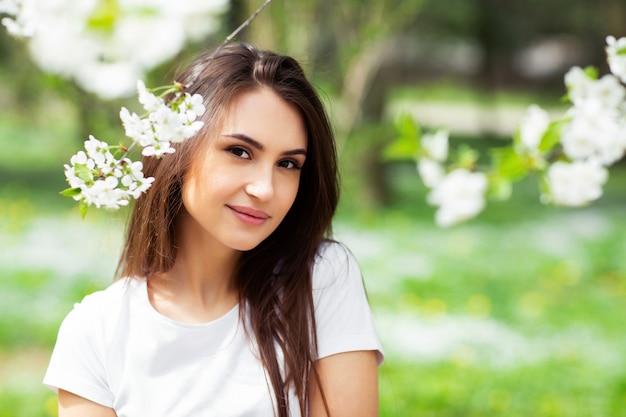 Muchacha que se coloca debajo de manzano floreciente en parque natural. Concepto de moda. Elegante joven en ropa de jeans disfrutando de jardín de flores en el soleado día de primavera. Maquillaje de belleza natural. Horizontal