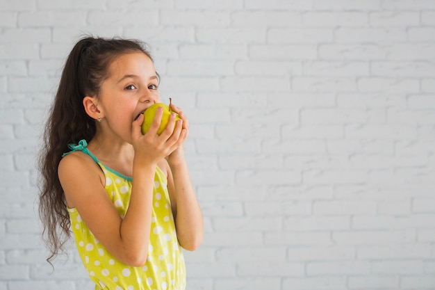 Foto muchacha que está parada delante de la pared que come la manzana verde