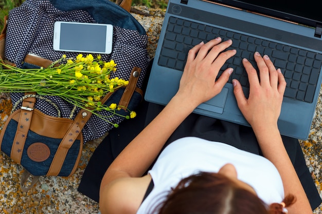 Muchacha que se sienta en hierba verde con las manos de la computadora portátil en el teclado.