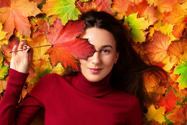 Muchacha que sostiene la hoja de arce roja disponible sobre fondo colorido de las hojas caidas.