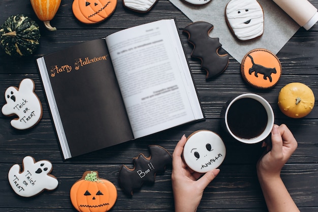 Muchacha que sostiene la taza de café, pan de jengibre para Halloween y que lee el libro de terror en un fondo de madera negro.