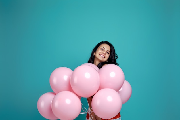 Muchacha rizada despreocupada sonriente en el vestido que sostiene los globos de aire del rosa en colores pastel aislados en fondo azul. Mujer joven feliz en una fiesta de cumpleaños. felicidad. espacio para texto