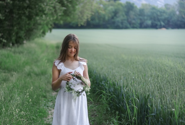 Muchacha romántica hermosa del preadolescente que hace la guirnalda verde y blanca de la cabeza de flores frescas al aire libre en el campo verde.