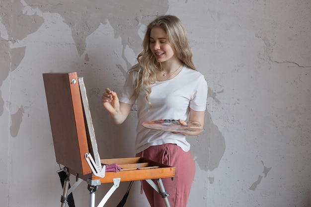 Muchacha rubia bonita sonriente de los jóvenes con el cepillo y la paleta que se colocan cerca de imagen del dibujo del caballete