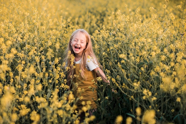 Muchacha rubia divertida del niño que presenta en el prado floreciente de las flores de la violación sobre el fondo de la naturaleza