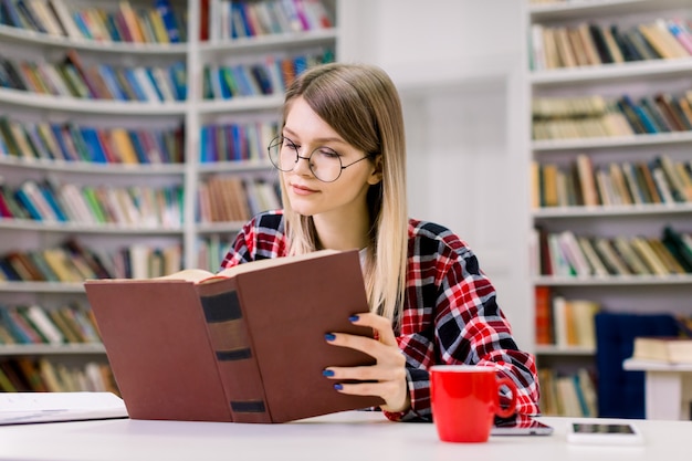 Muchacha rubia elegante atractiva joven del estudiante en las lentes que se sientan en la tabla, concentrada mientras que lee el libro en la biblioteca de universidad. Estantería con diferentes libros en el espacio