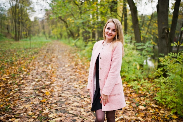 La muchacha rubia joven en la capa rosada presentó en parque del otoño.