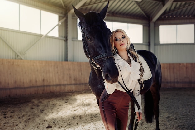 Muchacha rubia joven elegante hermosa que se coloca cerca de su competencia del uniforme de preparación del caballo