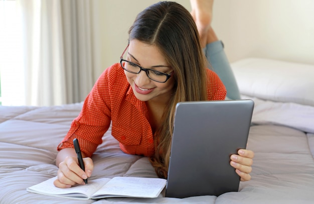 Muchacha sonriente del estudiante que lleva los vidrios que estudian en línea cómodo en casa.