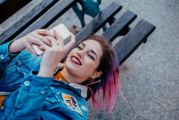 Muchacha sonriente con el pelo púrpura que miente en un banco, usando el teléfono.