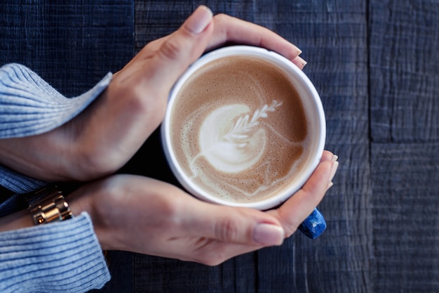 La muchacha sostiene una taza azul de capuchino en manos en una mesa de madera