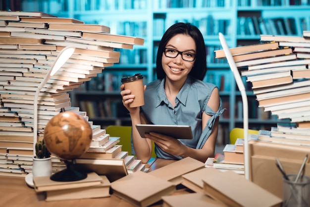 Muchacha con la tabla rodeada por los libros en biblioteca en la noche.