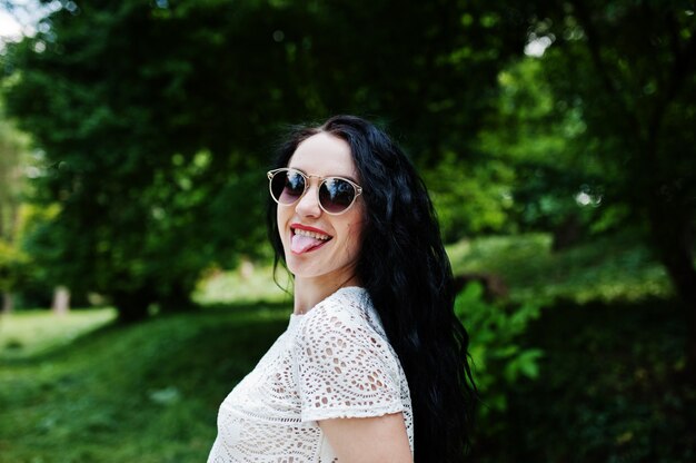 La muchacha triguena en la blusa blanca con las gafas de sol presentó en el parque.